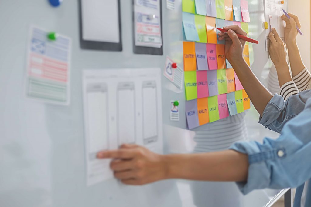 Programmer's Agile Experience. Na foto vemos um painel com várias anotações sobre o desenvolvimento de um produto digital e duas pessoas trabalhando nesse quadro.