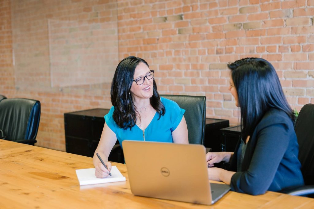 duas mulheres em uma sala de reunião sentadas a mesa. Uma com computador e a outra com um bloco de anotações falando sobre UX Design